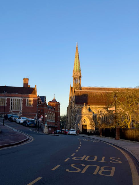 A narrow residential street viewed through an ornate, gothic-style stone archway with detailed carvings and pointy arch features. Beyond the arch, there are brick buildings, some with chimneys, and pa