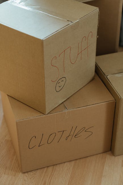 A woman with curly hair wearing a dark green t-shirt is sealing a cardboard moving box with packing tape in a well-lit room, possibly during a home relocation process. The box is placed on a wooden su